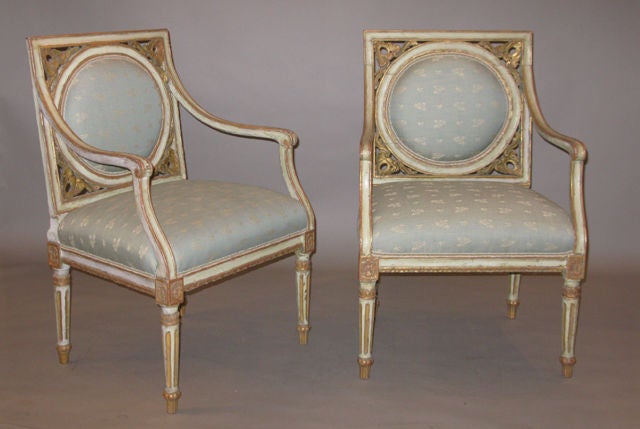 A pair of painted and gilded neoclassic arm chairs with a square back with a circular upholstered back within a painted and gilded border; surrounded by gilded and carved floral motif.  The apron with gilded rosettes above tapered legs with gilded