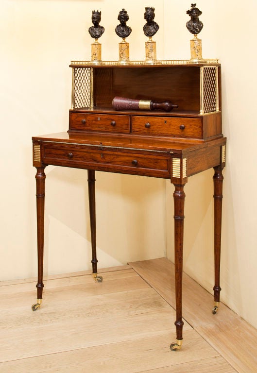 A rosewood and brass chiffonier desk having two open shelves over two drawers with a fold over writing surface over a drawer on turned legs with casters in the manner of John McLean, Circa 1810