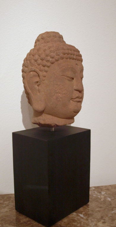 Dating back to the 18th century or earlier, this sandstone (reddish in color), Buddha head which is mounted to a bronze colored steel base, clearly makes an impressive statement. The entire head, front and back, including the headdress, is in