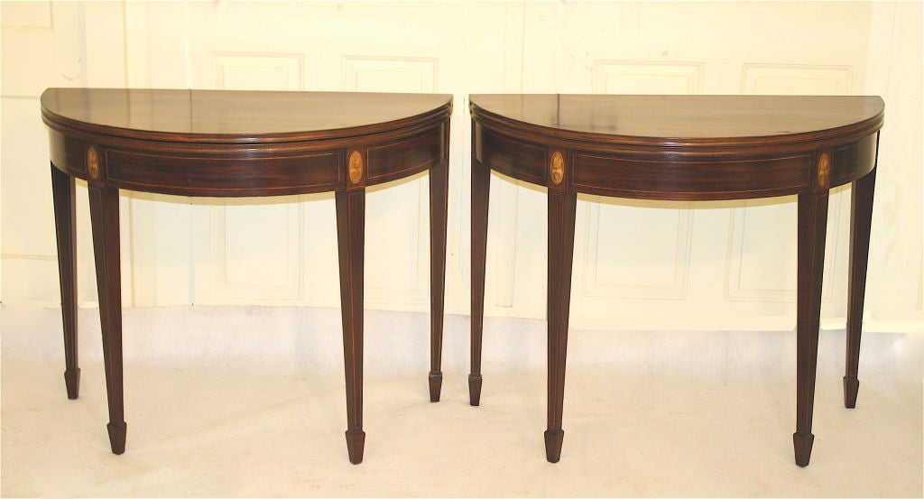 A perfectly matched simultaneously made pair of inlaid mahogany<br />
demilune form, Hepplewhite manner card/game tables.  Copious satinwood string inlays on the friezes and tapering legs ending in spade feet.  There are four oval foliate urn