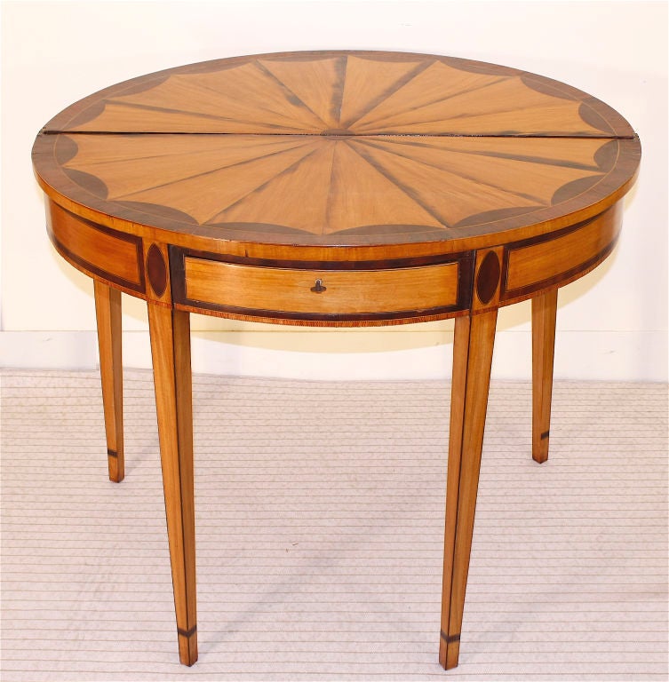 A 'specimen' intricately inlaid demilune card table, displaying the striking contrast of satinwood and rosewood; with a center drawer on tapering legs.  Double rear swing legs for stability and ease of seating; when desired.
