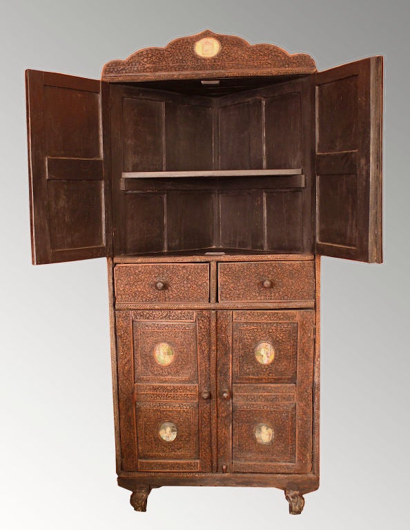 An intricately hand-carved corner cabinet decorated with nine oval reverse painted 'églomisé' plaques of Indian Brahmin, on a foliate vine case. Upper and lower shelf sections enclosed by pairs of panel doors; with two drawers in between.