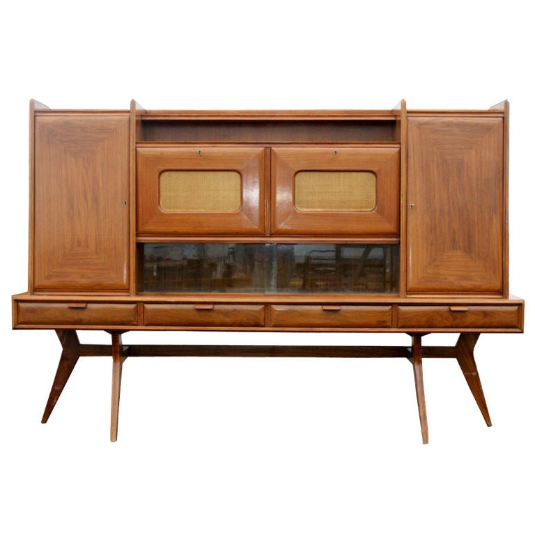 A mid century modern Italian wall unit in walnut featuring multiple storage and display areas.  Four drawers, including two partitioned for flatware.  Two drop front, mirrored dry bars.  Two doors concealing shelved storage.  Multiple display areas.