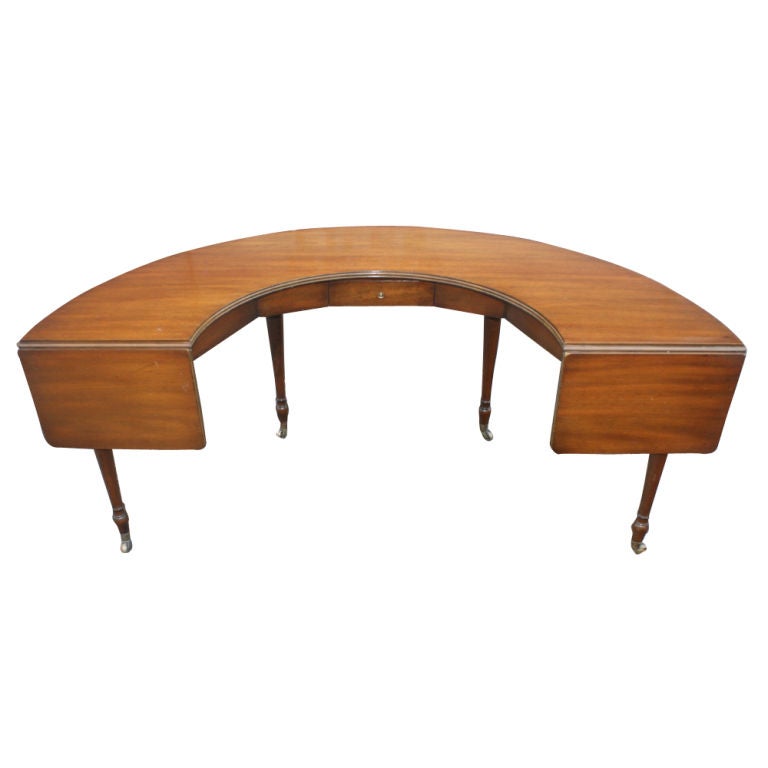 A Kittinger half-round writing desk in walnut raised on four legs with rounded spade feet on brass casters.  Two dropleaf extensions and one drawer.