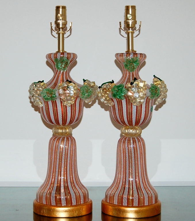 Matched pair of vintage Murano lamps, attributed to Dino Martens. Various shades of ORANGE striped glass are adorned by Murano glass fruit. The two pieces of filigrana glass are cinched at the waist by a doughnut full of gold.

The lamps are 24