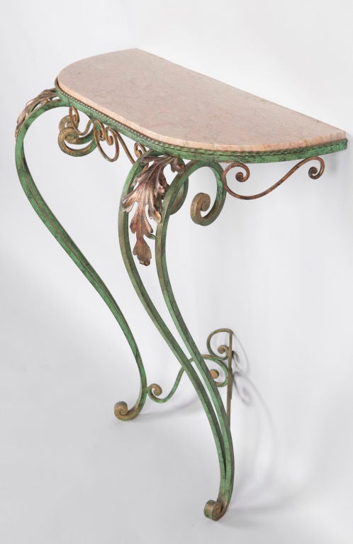This French Demi-Lune Wall Console Table has a forged iron base with a green and gold patina, with motifs of scrolls and acanthus leaves.  The marble top has shades of gray and pink.