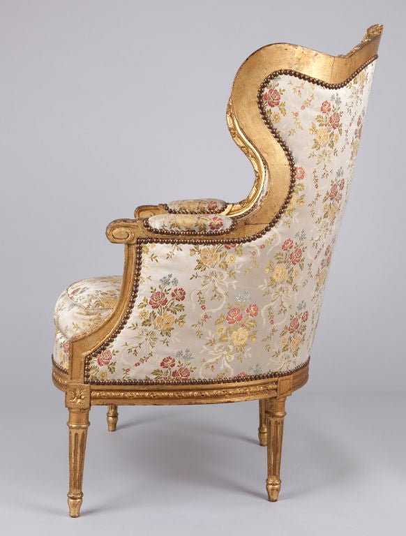 This fabulous Louis XVI Style Giltwood Bergere Armchair is upholstered in silk with floral motifs.  It has a high back with wings and a carved cresting with flowers. The frame has padded armrests and carvings of acanthus leaves, ribbons and