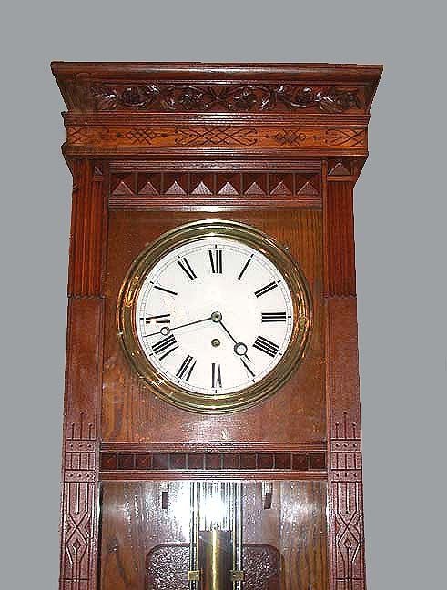 Classic Gilbert #12 regulator in Eastlake oak long case with floral and foliate carved band running under cornice and over a row of incised decoration; with brass ringed clock face flanked by square columns with fluted terminals. The glass fronted