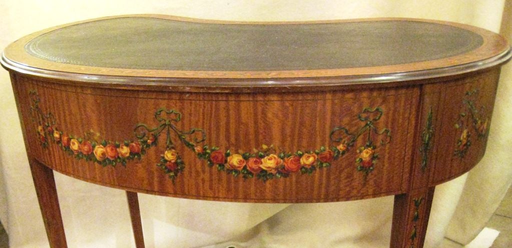 Edwardian satinwood kidney form desk with conforming inset leather top surrounded by painted floral garlands, over three drawers each with crossbanded borders around garlands and original bail handles, raised on tapered legs on brass casters.