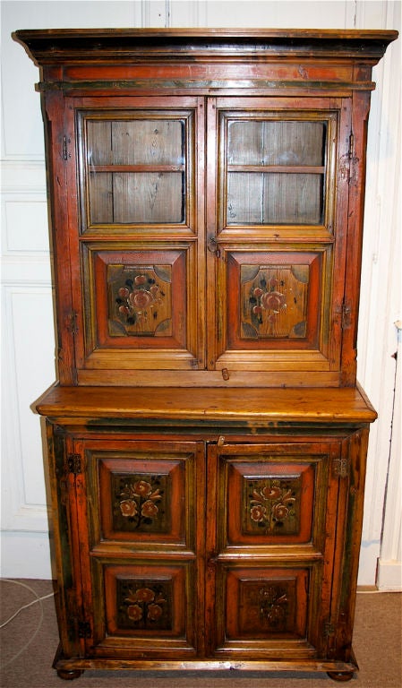 This country cupboard from the Baltic is made of pine and retains its original worn paint decoration. There are fielded panel doors with its original flower motifs while the top has two panels of glass. The size is unusually small.