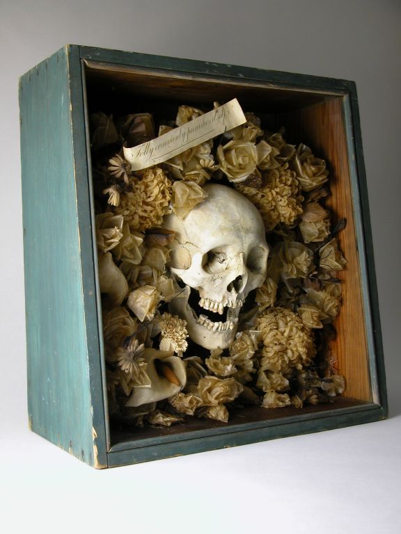 A Temperance movement glass enclosed painted pine shadow box with canted front containing a human skull wreathed in waxed paper flowers.  <br />
<br />
Epitaph rendered on a ribbon above: 