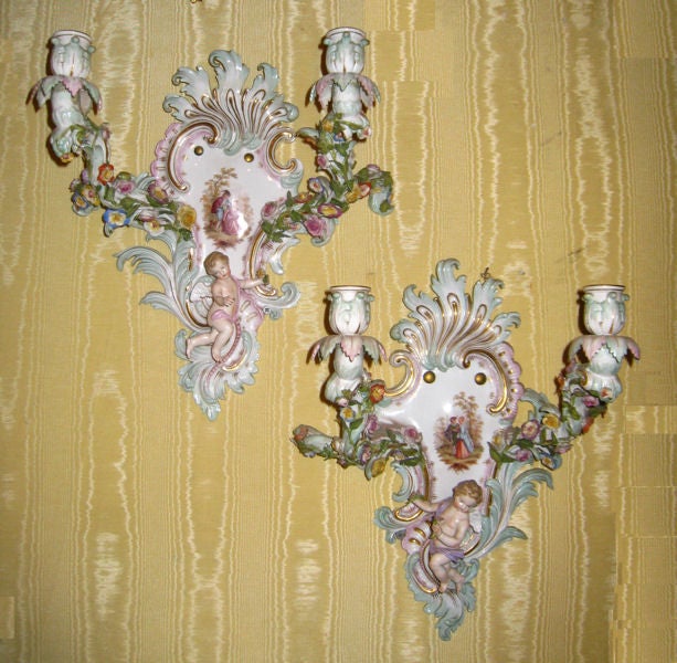 This pair of porcelain two-candle sconces with rocaille-cartouche form by Meissen date from the nineteenth century. They feature foliate nozels and drip pans at the end of floral-encrusted branches, with three-dimensional cherub figures and