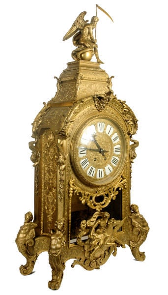 This mantle clock in the French Louis XIV style features elaborate ormolu bronze decoration. The face is decorated with enamelled porcelain numbers, surmounted by a Greek mask. The original key and pendulum with the Sun King emblem are present.
