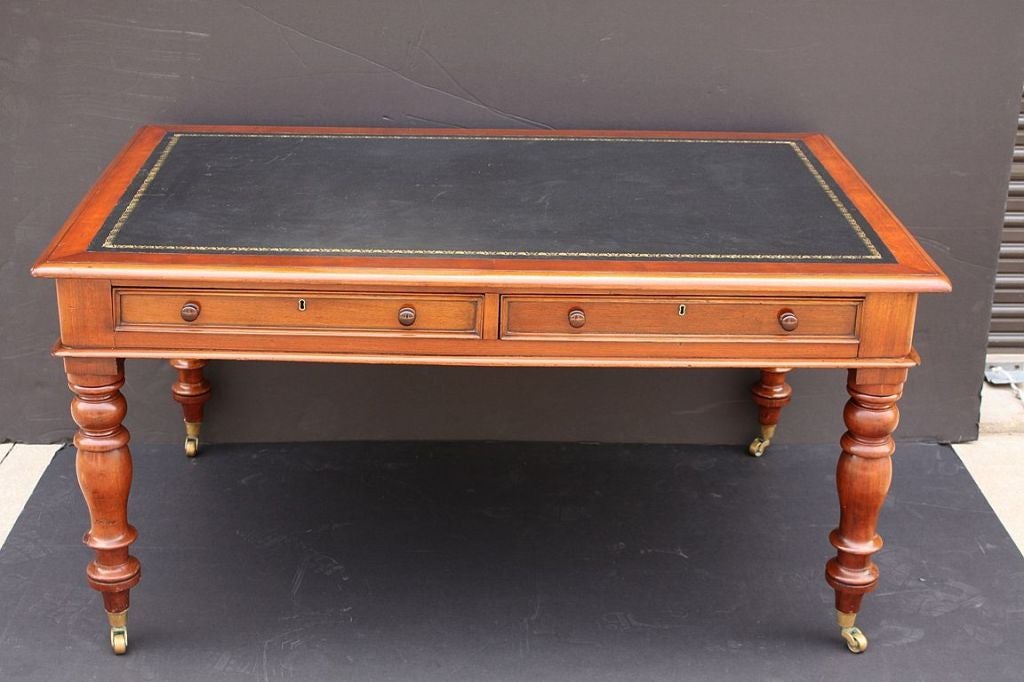 An English partner’s writing or library table of mahogany,<br />
featuring a black leather top embossed with gilt design, <br />
over two long drawers on opposing sides,<br />
resting on turned baluster legs with brass casters.<br />
<br