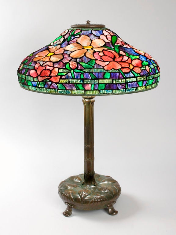 A Tiffany Studios New York glass and bronze “Peony” table lamp, featuring a leaded glass shade in mottled hues of purples and blues, decorated with multi-colored peony blossoms. The shade is further accented with three lower rows of mottled green