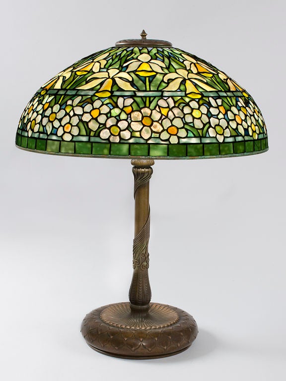 A Tiffany Studios New York “Jonquil-Daffodil” leaded glass and bronze table lamp, featuring orange/yellow jonquils in the upper portion and white petaled  flowers with orange/yellow centers in the lower portion, all resting on a green leafed ground.