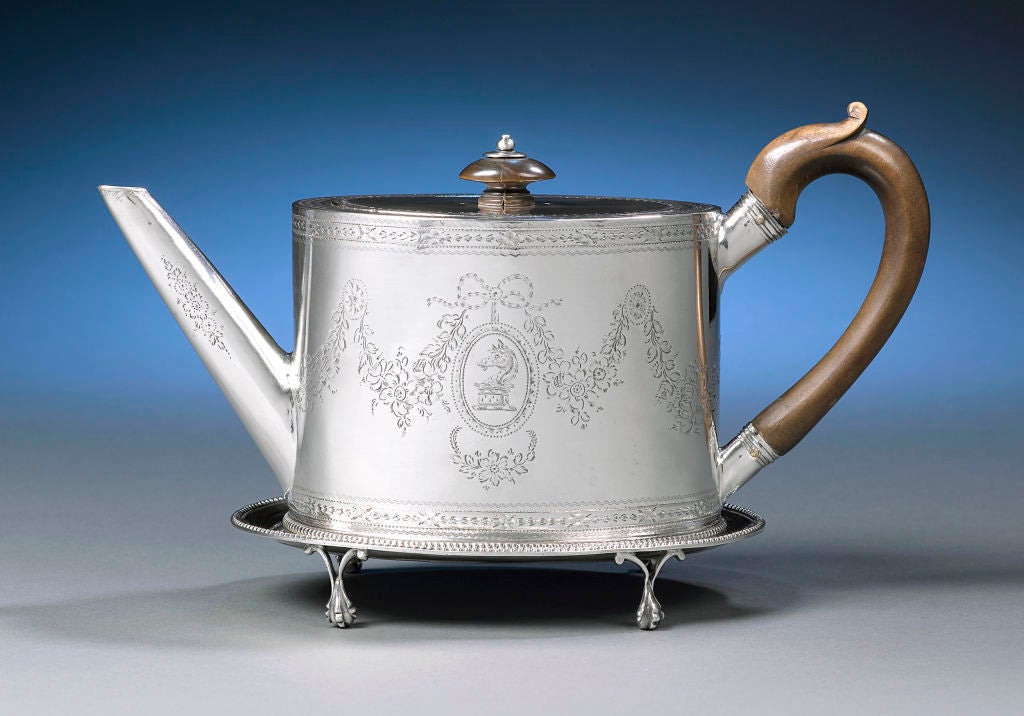 This extraordinary George III-period silver teapot by Hester Bateman not only displays this master silversmith’s signature style, but is also accompanied by a rare silver stand. This charming teapot, with its oval, straight-bodied shape and wooden