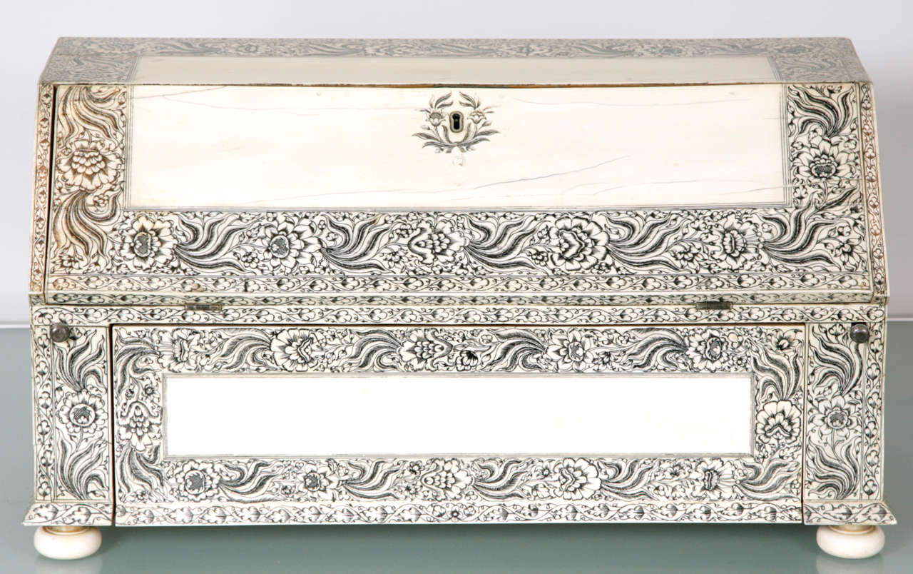 The outside entirely veneered in ivory engraved with borders of composite flowers emanating from tufts of 4 wavy banana leaves, the hinged front opening to reveal three central pigeon holes flanked on either side by three curved ivory faced drawers,