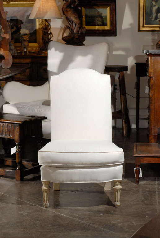 A 20th century painted French single slipper chair featuring a high, straight rectangular backrest with a crossbow-shaped top, covered with a smooth white muslin upholstery. The seat also features a loose fitting trapezoidal cushion. This armless