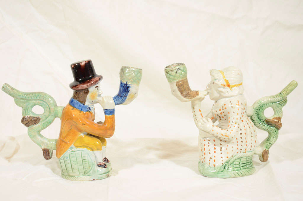 A pair of Prattware pipes modeled in the form of Martha Gunn and a sailor. Each is  seated on a green washed base. The bowls of both pipes are modeled as a man's head, the stem and mouthpiece are modeled as 2 dolphins.
  Martha Gunn worked as a