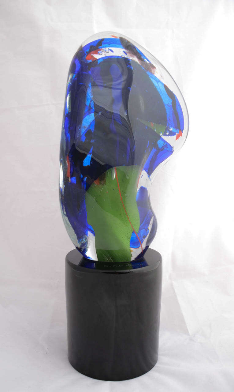 This abstract piece is a single curved mass of clear glass, internally decorated with swirls of blue, red and green and bubbles of air. It is on a cylindrical hollow black glass base which is etched with the signature S.Toso, Murano, for Stephano