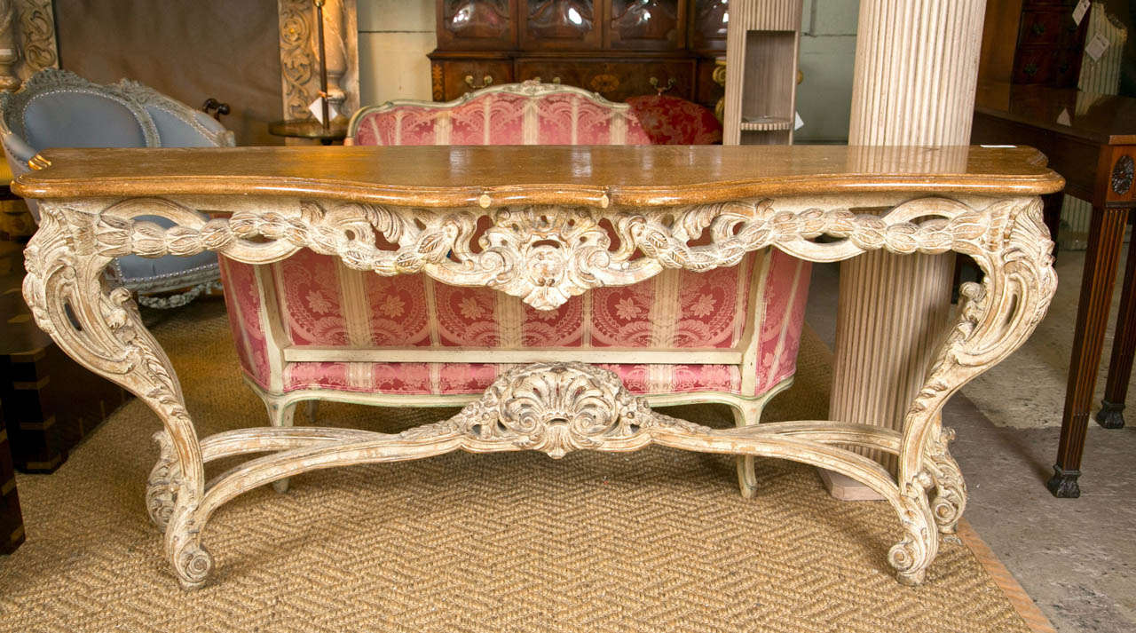 French Rococo style console table, the serpentine shaped wood top over a distress-painted base decorated with scrolls and shell carvings, joint by a stretcher and raised on scrolled feet. Manner of Maison Jansen