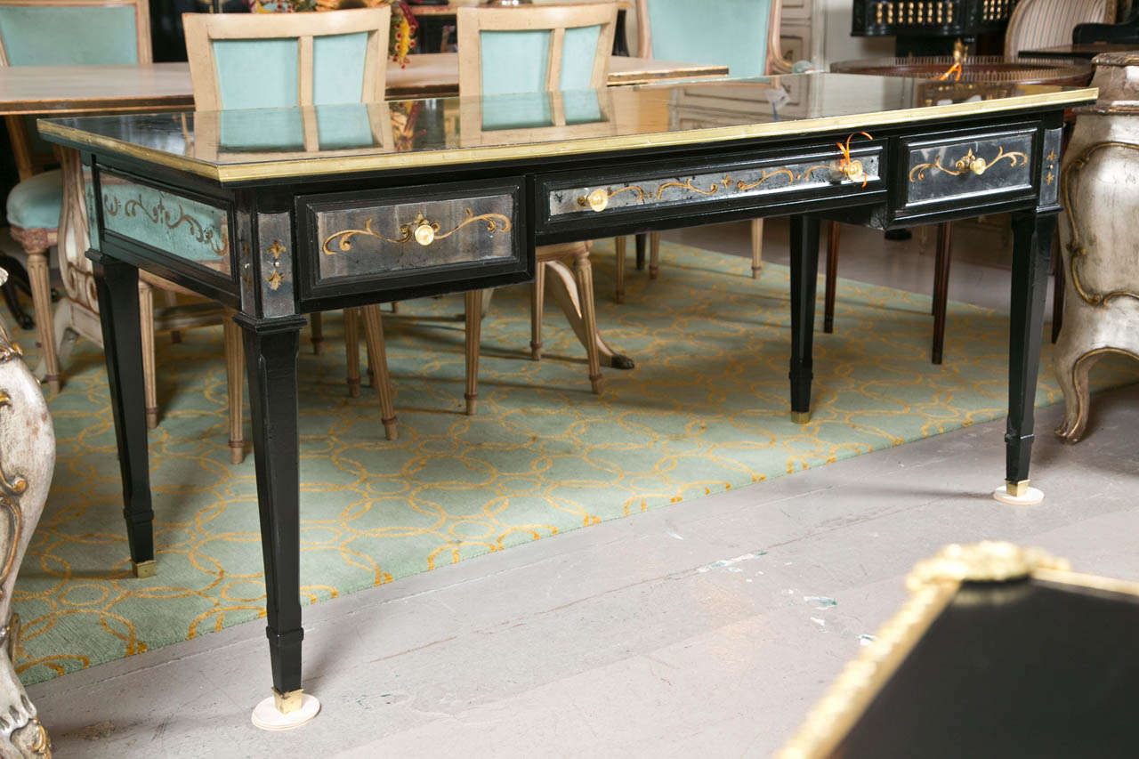 A glamorous French Louis XVI style desk, overall ebonized, the bronze banded top with verre eglomise glass veneer, over a narrow frieze fitted with three drawers and decorated with eglomise glass panels. Raised on squared tapering legs ending in