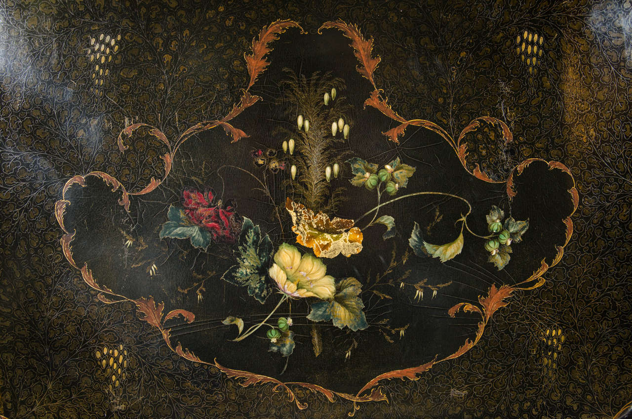 A black lacquered papier mâché tray with flowers and leaves subtly painted in soft colors with a chinoiserie motif decorated overall with gold accents depicting long tailed birds flying above elegant mandarins in a flower filled garden.