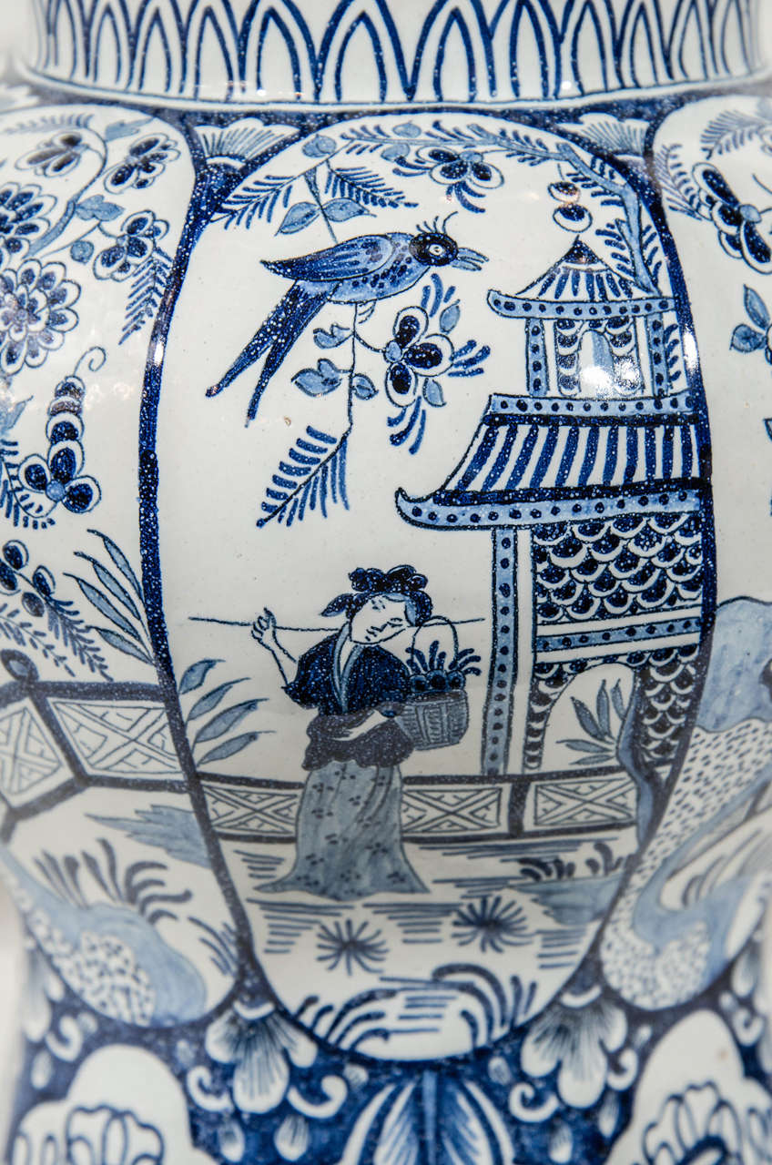 A pair of Dutch delft vases painted in blue and white with scenes in panels. In one set of panels a young woman holding flowers stands before a pagoda. The next set of panels is painted with a flower filled garden.
The cover and base of each vase