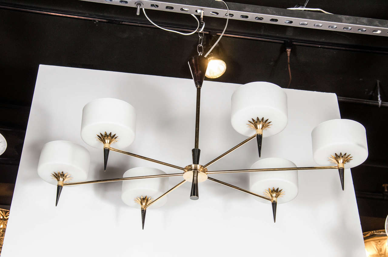 This very sophisticated Art Deco chandelier in the manner of Adnet features an elongated elliptical design with six arms; each arm extends out to support a cylindrical frosted glass shade on a torch-like decorative spoke with brass starburst detail.