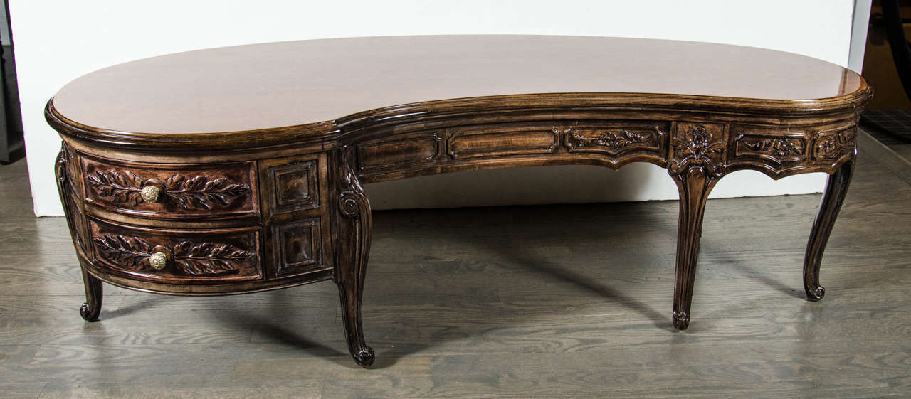 This organically shaped 1940's walnut Hollywood Regency cocktail features elaborate hand carved laurel and geometric styling. It offers a pair of drawers at one end, as well as stylized floral pulls in antique brass and cabriole legs with Flemish