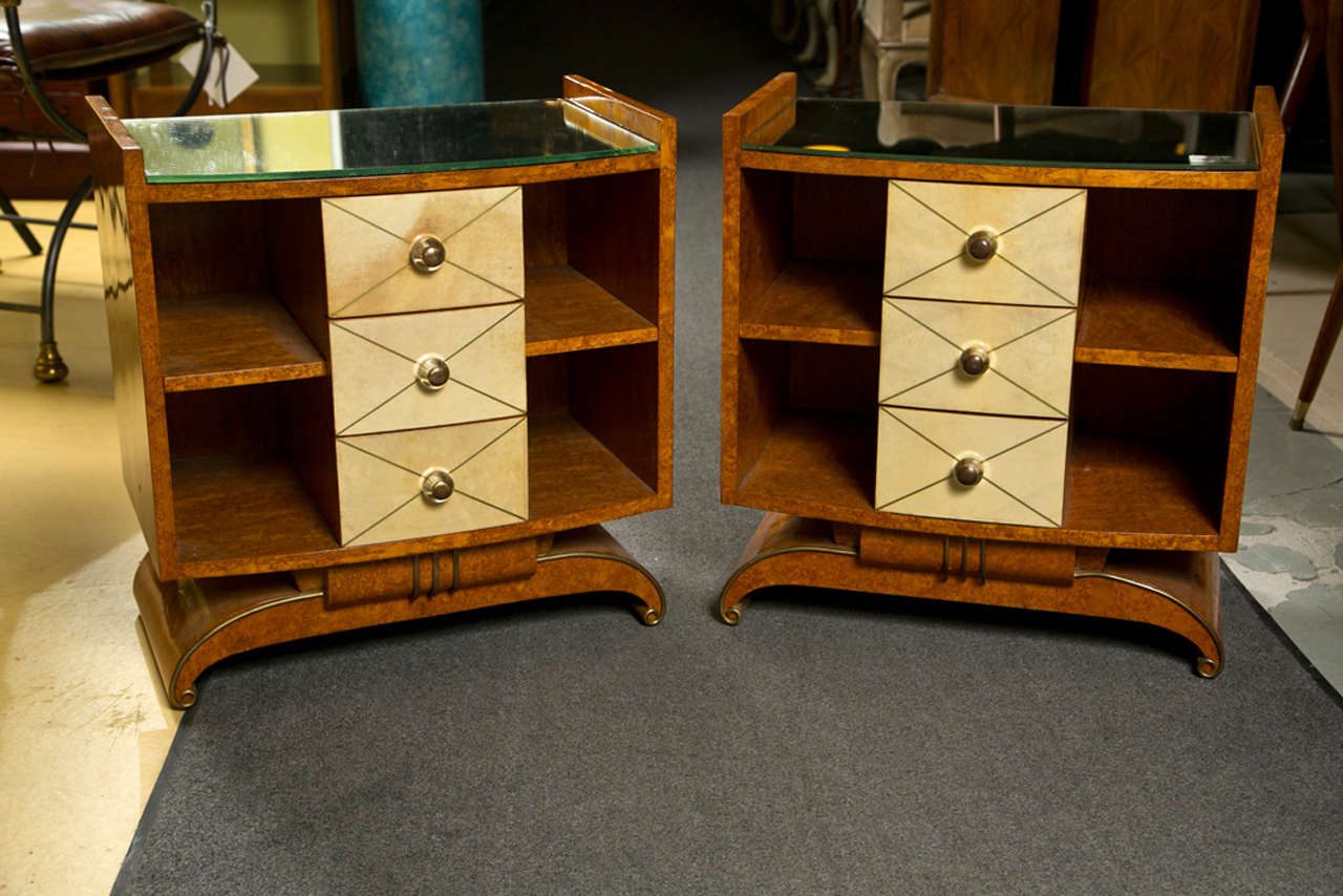 Pair of Art Deco style burlwood bedside tables, circa 1950s, each has glass top, atop a set of three drawers with parchmont veneer and lucite knobs flanked by shelving storage each side, raised on a dome-form plinth. 