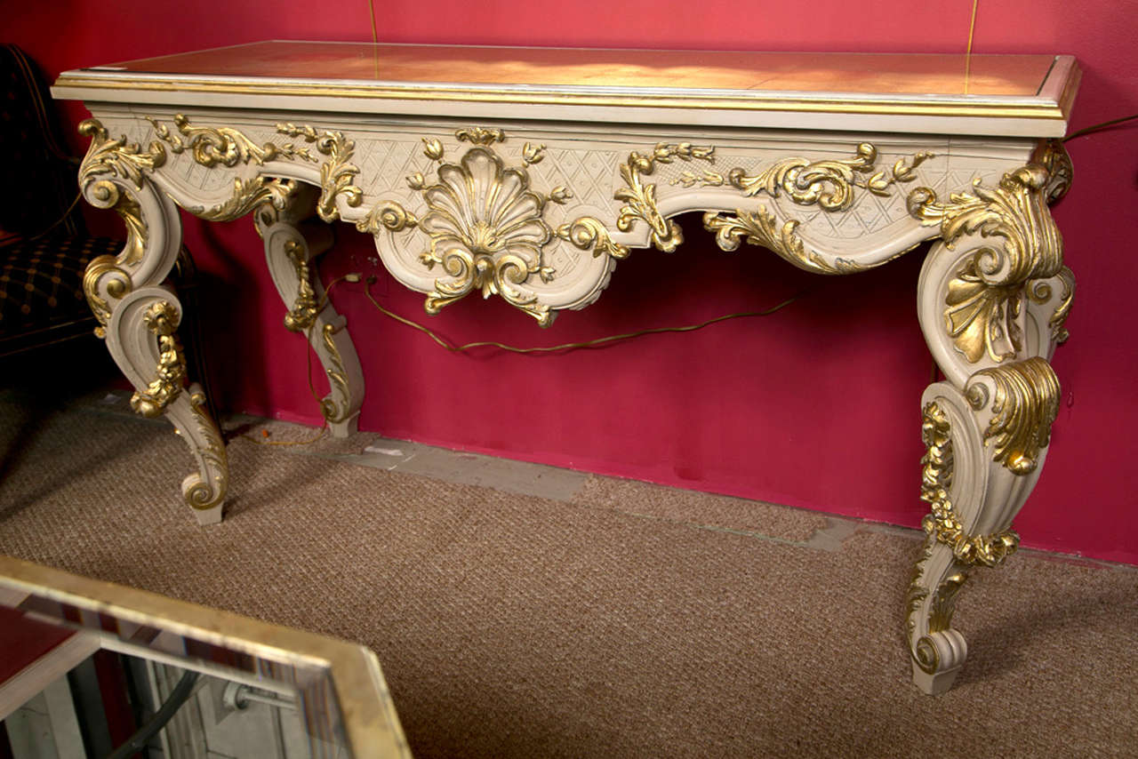 A French Rococo style crème peinte and parcel-gilt console table, circa 1940s, the shaped top with gilt-glass insert, over a scallop-shaped frieze decorated with intricate carving of scrolls and shell motif, supported by like-design scrolled legs.