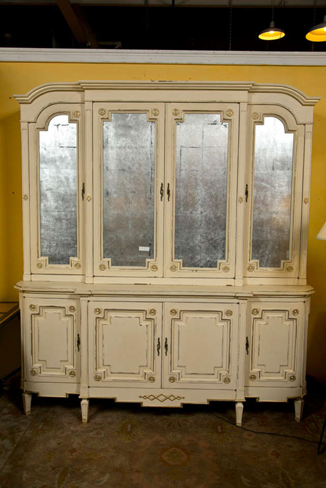 A charming ivory distress-painted gilt gold decorated bookcase, circa 1950s, the molded top over a four-door cabinet, each door with silver-leaf glass opening to shelving paper decorated interior, atop a conforming cabinet raised on short fluted