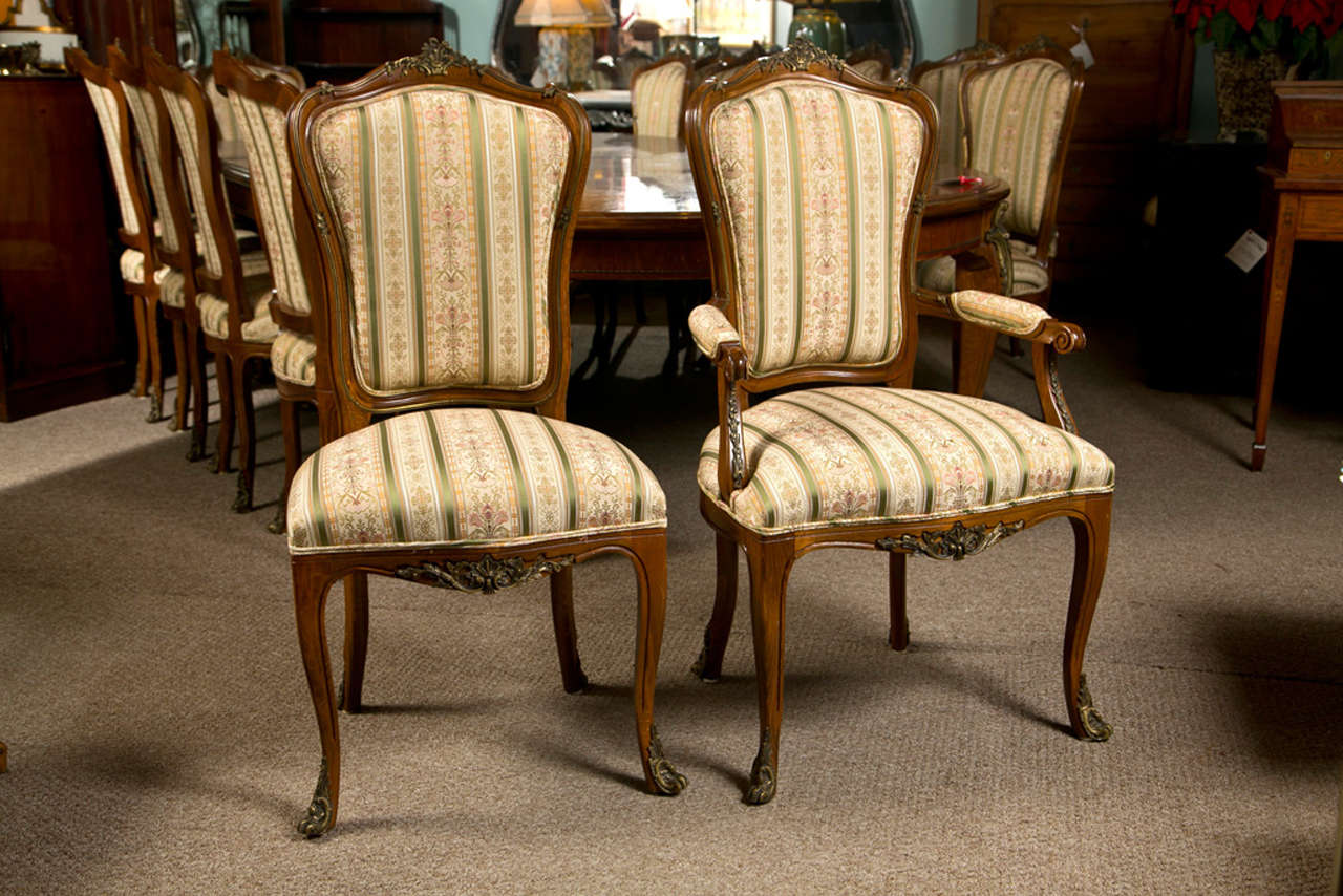 A group of six French Louis XV style dining room chairs. Each chair with a set of front and rear bronze sabots as well as a central bronze apron decoration. The backs all with bronze framed upholstered back rests all adorned with a bronze top