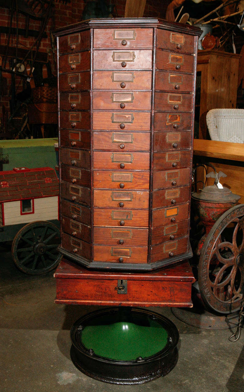 This Hexagonal Cabinet Stands on a square base sits on top of a circular painted metal revolving stand. This general store relic has 96 triangular drawers with wooden knobs and metal label plates that contain some remnants of paper labels. The