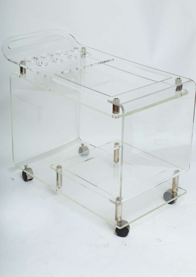 A practical bar cart in lucite on wheels with glass holders shelf and drinks shelf on chromed columns . Comes with glasses and additional wet tray.