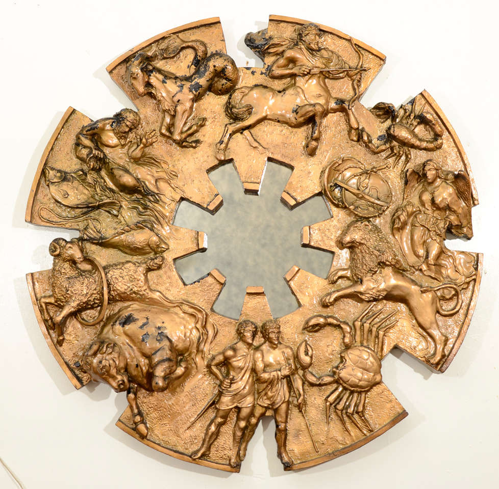 Wall mirror framed by a bold zodiac wheel with the twelve astrological signs. The frame is deeply sculpted resin with a bronze metallic finish. The mirror has a mottled smokey effect. 