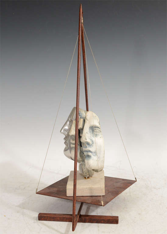 An abstract sculpture depicting three different views of a face. The piece sits on a platform suspended from a triangular metal 