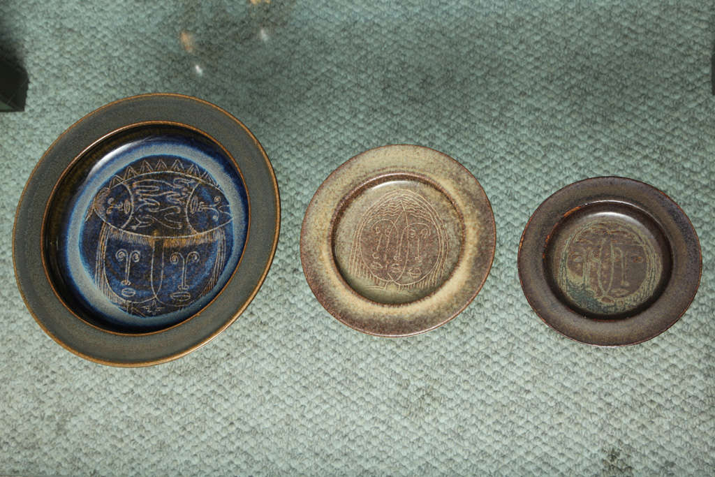 Three wide-rimmed bowls made of earthenware, in browns, creams and blues. Each bowl a similar form in concentric sizes and each with face decorations inside. All three pieces are signed and two are dated from the 1950s. Diameters for the three