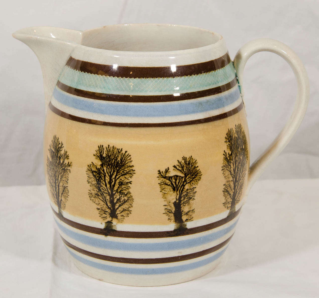 A large Mocha ware jug  decorated with machine turned bands of slip in light tones of blue and green and thinner bands of dark brown framing a single broad band of beige which has within a dendritic design resembling a row of trees at the edge of a