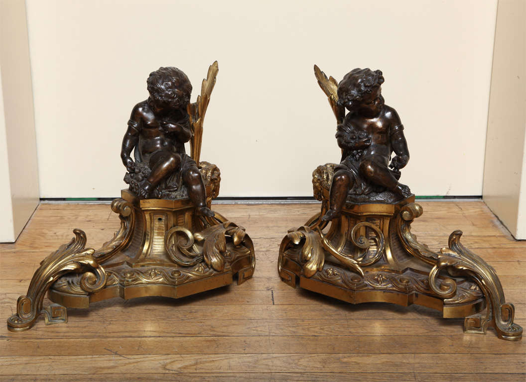A pair of French bronze chenets with patinated bronze putti vesting leaves and scrolls.