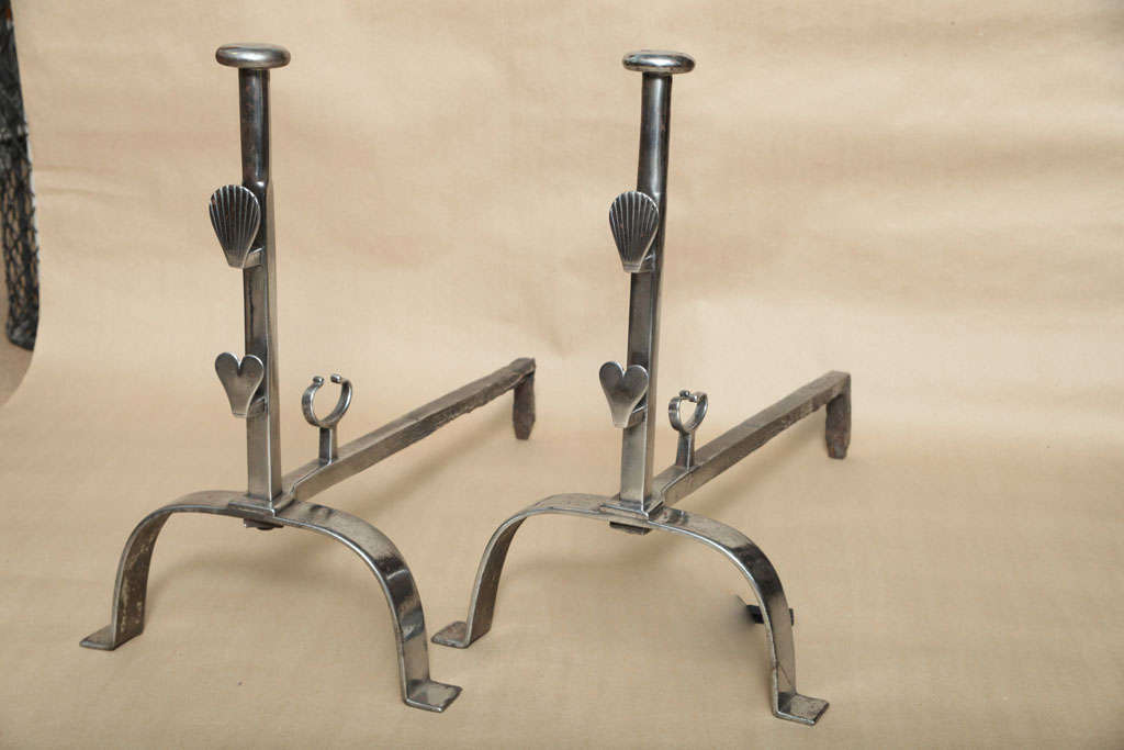A fine pair of 18th century English andirons made by a local blacksmith in the folk-art manner with heart and clamshell spit supports over arched feet with shaped clips to back shafts. In a handsome polished steel finish.