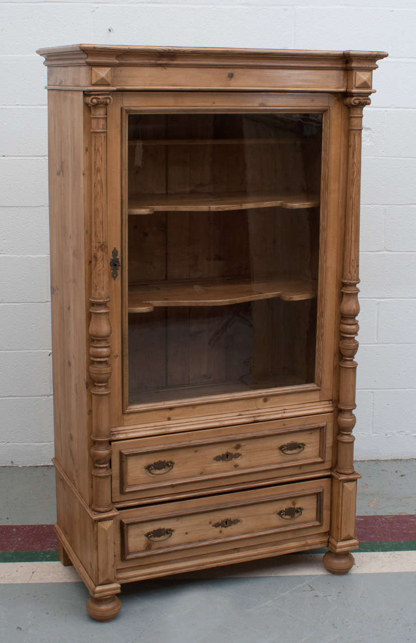 A beautiful display cabinet featuring a single door with original glass, scalloped and morticed shelves to the interior and two hand-cut dovetailed drawers, all flanked by two turned columns. All original except replaced bun feet.