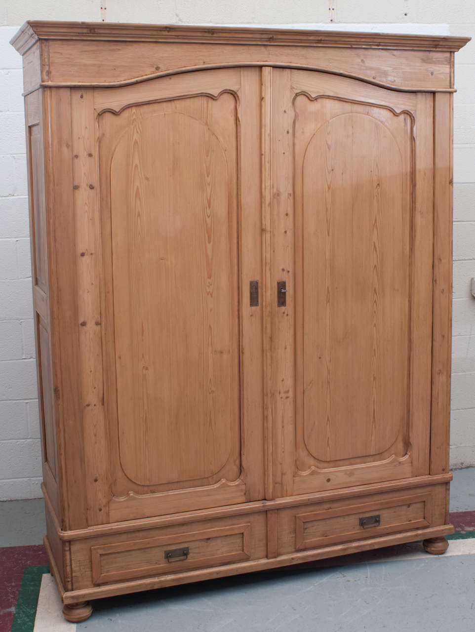 A large and handsome pine armoire featuring raised panelled sides, two scallop panelled arched doors with two faux drawer fronts to the base. Original wooden hanging pegs inside. Replaced crown molding. This piece completely breaks down for ease in