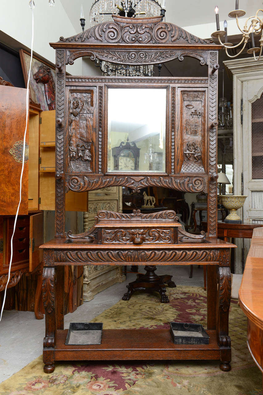 This is a very nice heavily carved oak hall stand with the original beveled mirror. To the top either side it has a scene with carved people, and to the glove box there is a carved lions head. We have the drip trays to the base. This is in excellent