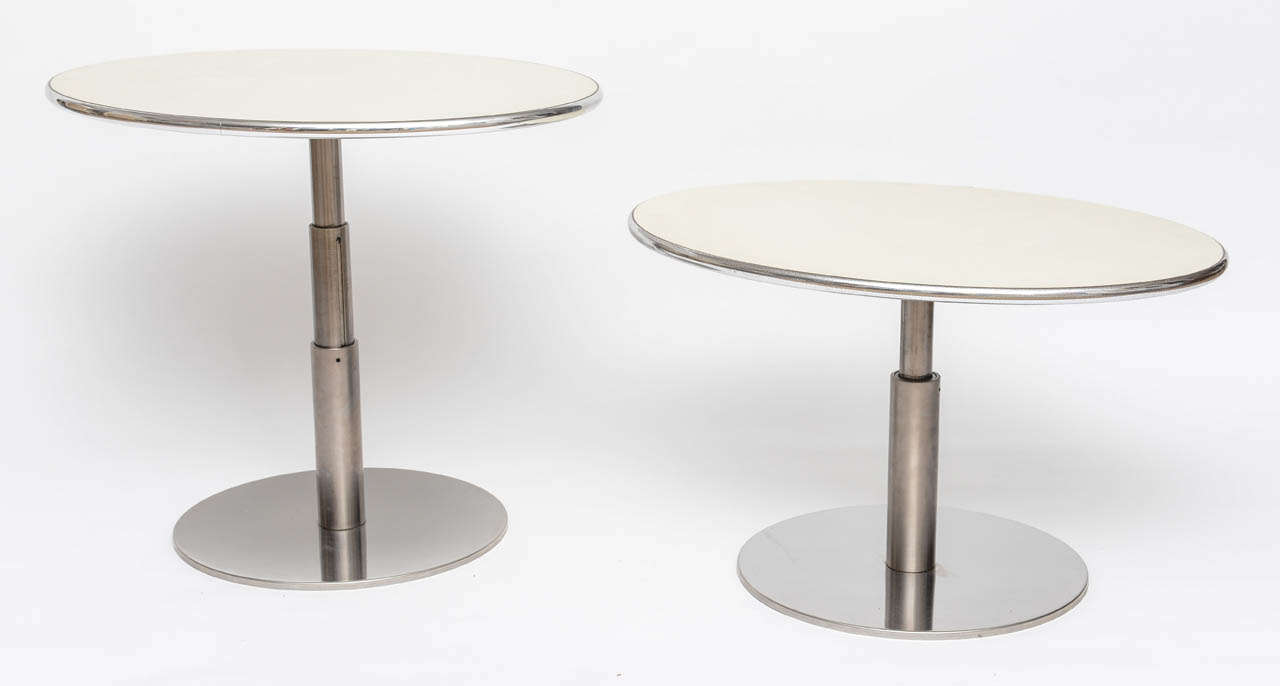 Wonderful pair of side tables with off white oval laminate tops banded in chrome. The round stainless steel bases support the three height pivoting center pole which locks the table tops in place at each of the three varying heights; very low,