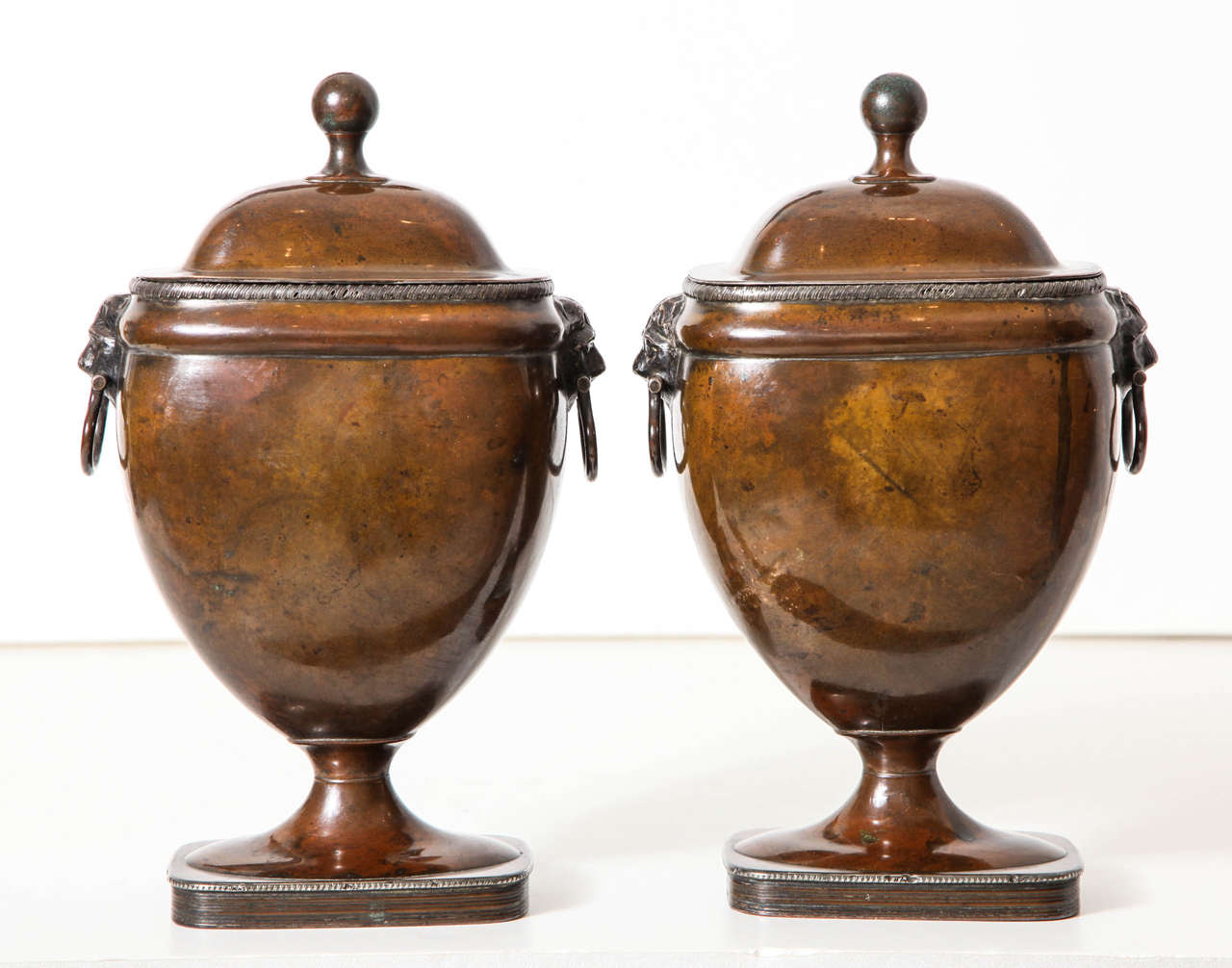 A pair of 19th century English (formerly) sheffield-plated covered urns with domed lids and ringed lion's masks on rectangular bases, circa 1830 / The worn surface has lost its original silvering rendering the bodies and lids with a wonderfully