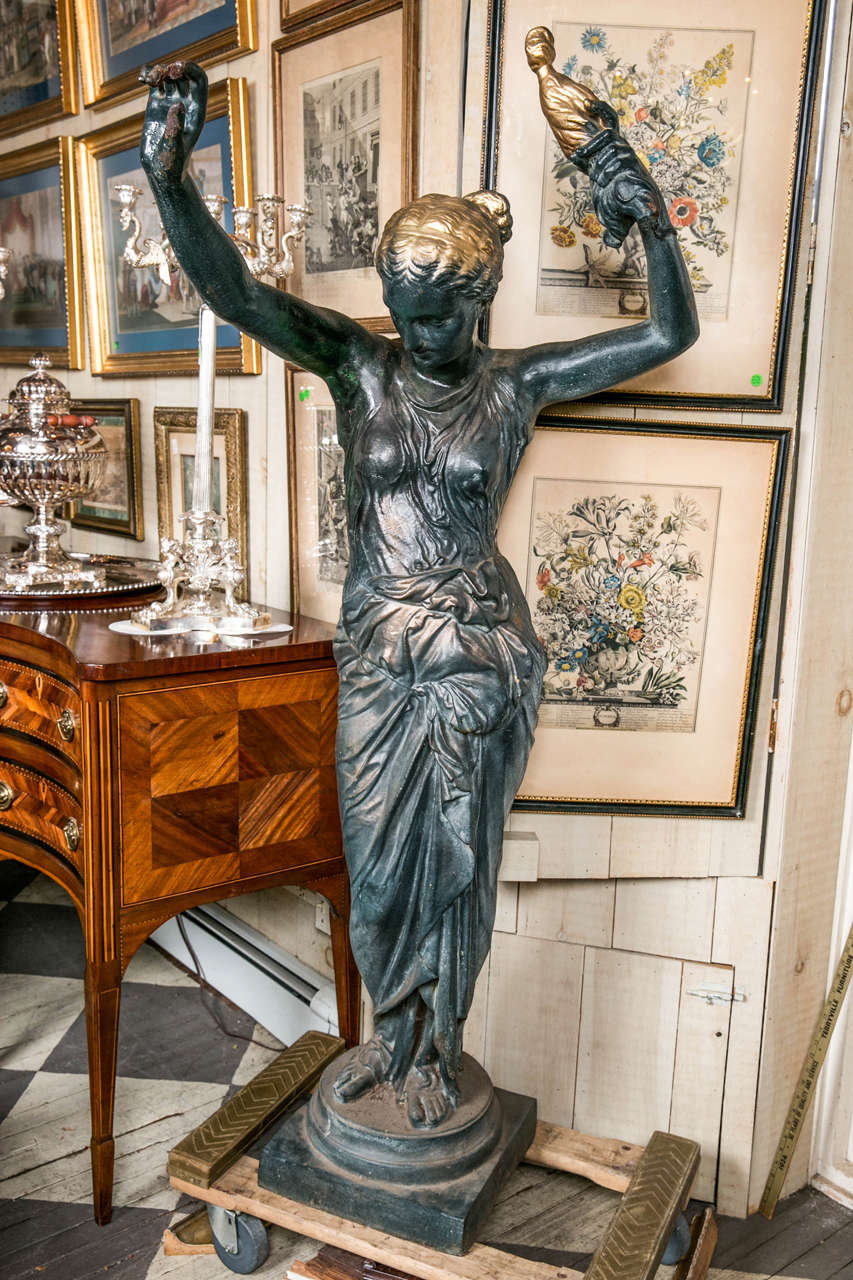 This figure of cast iron is done in the Greco Roman style. Her left hand holds aloft a flaming torch. Her right hand, also aloft, is empty and projects approximately 32 inches.
She wears a flowing draped gown in the Greco Roman style. Her bare feet