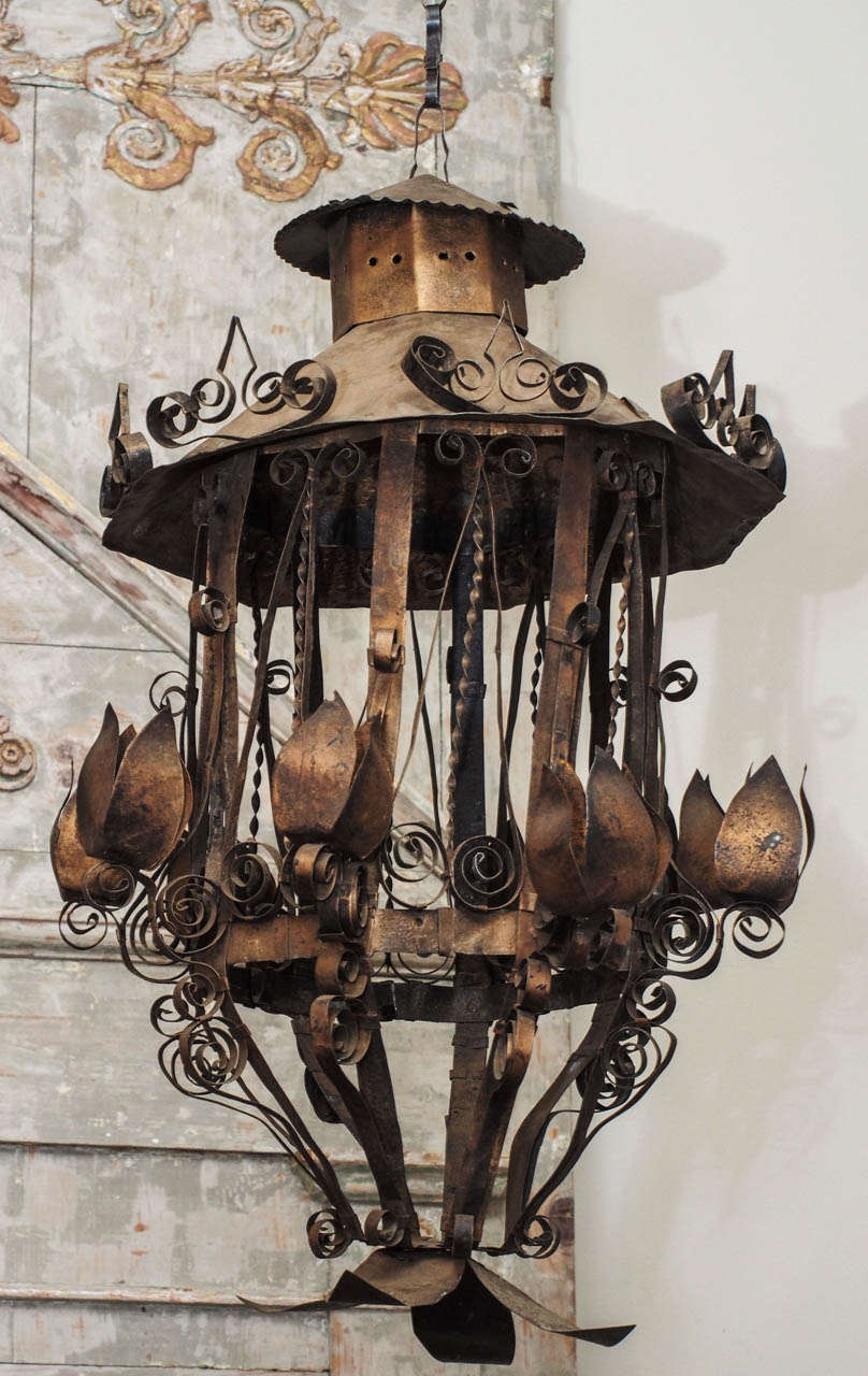 A whimsical tole ceiling light, the central support in the form of a lantern, capped with a conical top with a pierced chimney, the frame supporting seven  arms ending in tulip shaped candleholders. Overall decorated with scrolling metalwork, and